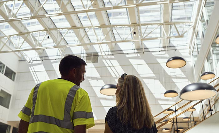 Ein Facharbeiter und eine Kollegin betrachten die Installation eines Oberlichts vom Inneren eines sanierten Gebäudes aus.